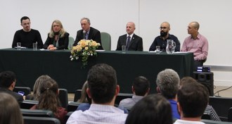 Em primeiro plano, pessoas sentadas de costas olhando para a mesa de honra. Na mesa de honra, seis pessoas estão sentadas lado a lado