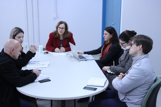 Foto aberta mostra seis pessoas sentadas ao redor de uma mesa oval