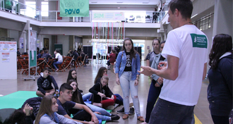 Estudante com a camiseta da UFFS em pé, explicando sobre a Instituição para jovens que estão sentados em um tatame, no hall do Bloco A