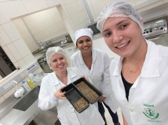 Três mulheres vestidas com jalecos apresentando um alimento.