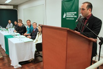 Um homem falando para o público. Ao seu lado, integrantes da mesa de honra observam.