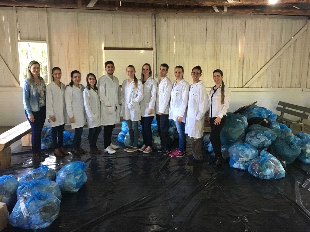 Onze pessoas lado a lado, olhando para a câmera, em um espaço onde contém sacos com os resíduos sólidos recolhidos.