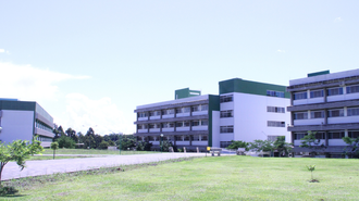 Foto em plano aberto da UFFS - Campus Chapecó. Em primeiro plano, um gramado e uma grande calçada. Aprecem, ao fundo, parte de três dos prédios do campus; todos pintados de branco e verde.