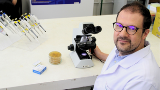 Professor está sentado em frente a uma bancada, de jaleco, com um microscópio e outros instrumentos de laboratório