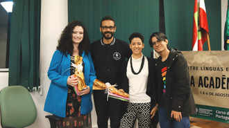 Na foto aparecem quatro pessoas em pé, em um auditório. Duas mulheres seguram presentes