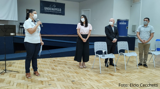 Foto com quatro pessoas em pé em um auditório, duas mulheres à esquerda e dois homens à direita. A mulher mais à esquerda está falando ao microfone.