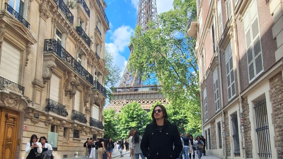 Imagem de um jovem em uma rua. Ao redor, prédios antigos, e, ao fundo, árvores e a Torre Eiffel