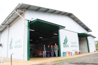 Foto aberta do galpão da área experimental, uma estrutura pintada de branco por fora. Em frente à estrutura estão três homens e uma mulher: professor Roberto, Diego, Luciano e professora Crhis