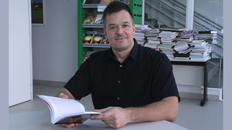 Foto de um homem branco, sentado em frente a uma mesa, com as duas mãos segurando um livro abeto.