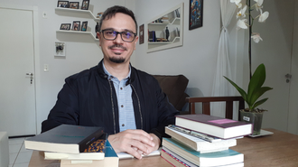 Na foto, Allan está sentado em frente a uma mesa com livros.