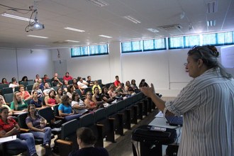 Foto em plano aberto mostra a professora Noeli em primeiro plano, falando e gesticulando. Em segundo plano, o auditório com os professores sentados