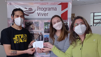 Em frente a um banner do Programa Viver, jovem presidente do CA entrega envelope a duas mulheres. De máscara, todos olham para a câmera e é possível perceber que estão sorrindo.