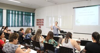 Uma professora fala e gesticula na frente de outras pessoas que estão sentadas em cadeiras, em uma sala de aula