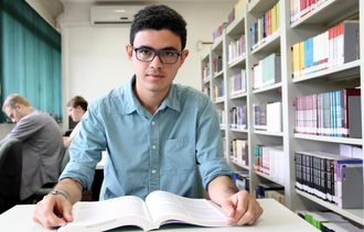 Professor está na biblioteca, sentado em uma cadeira, de frente para uma mesa, com um livro aberto em sua frente