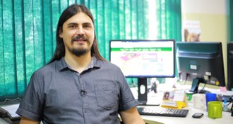 Foto com enquadramento fechado no rosto do professor, que está sorridente, sentando em frente a uma mesa. Em segundo plano há uma mesa, com um teclado e um monitor de computador