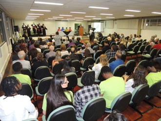 Foto da colação de grau com a plateia sentada e formandos no palco fazendo o juramento