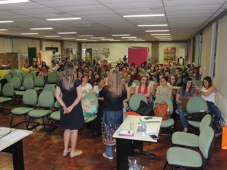 Duas professoras de pé, falando para alunos que estão sentados na plateia