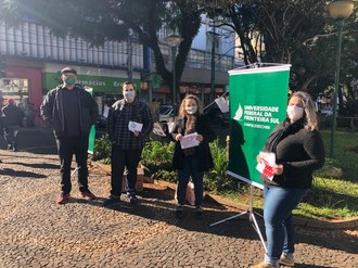 Foto de quatro pessoas de pé, em uma praça, usando máscaras, com um banner da universidade próximo a elas