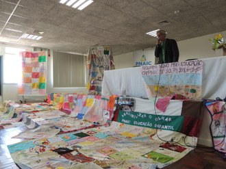 Foto com o palestrante ao microfone, atrás de uma mesa, e, na frente, ao chão, trabalhos em tecido feitos pelos participantes da formação