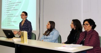 Foto com três mulheres sentadas e uma de pé, esta falando e gesticulando