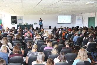 Professora falando ao microfone para uma plateia que está sentada