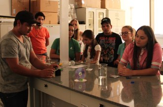 Na imagem oito estudantes estão poscionados ao lado de bancada de laboratório onde realizam experimentos.