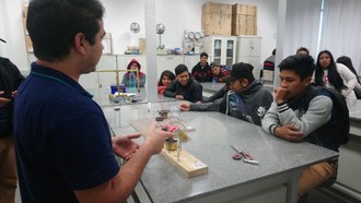 Dentro de um laboratório, estudantes acompanham demonstração de experimento da área de Física que está em uma das bancadas do laboratório. Um dos estudantes apresenta aos demais informações sobre o experimento.