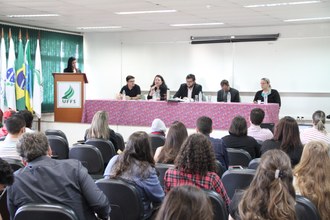 Plateia em auditório oberva pessoas no palco formando mesa diretiva. Uma pessoa faz pronunciamento.