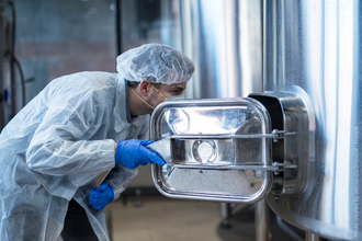 Foto mostra um homem usando uniforme de proteção com rede para cabelo, luvas e máscara. O homem abre uma máquina na linha de produção de uma fábrica de alimentos e observa o interior do equipamento.