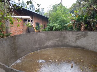 Foto mostra parte de uma cisterna. Um cano de pvc direciona a água para o centro da cisterna. Ao redor da estrutura estão diversas plantas e uma construção com tijolos a vista.