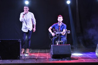 Na imagem duas pessoas estão no palco cantando. Um homem está em pé com o microfone na mão e outro está sentado, tocando violão.