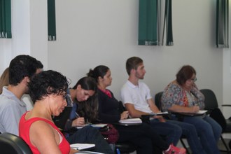 Grupo de pessoas sentadas, algumas olham para frente enquanto outras fazem anotações durante palestra.