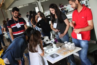 Na imagem estudante observa experimento em microscópio. Ao seu redor outros estudantes acompanham apresentação.