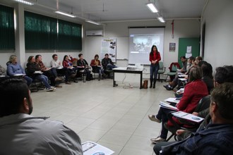 Na imagem pessoas sentadas forma um círculo em sala de aula. A frente da plateia professora faz apresentação.