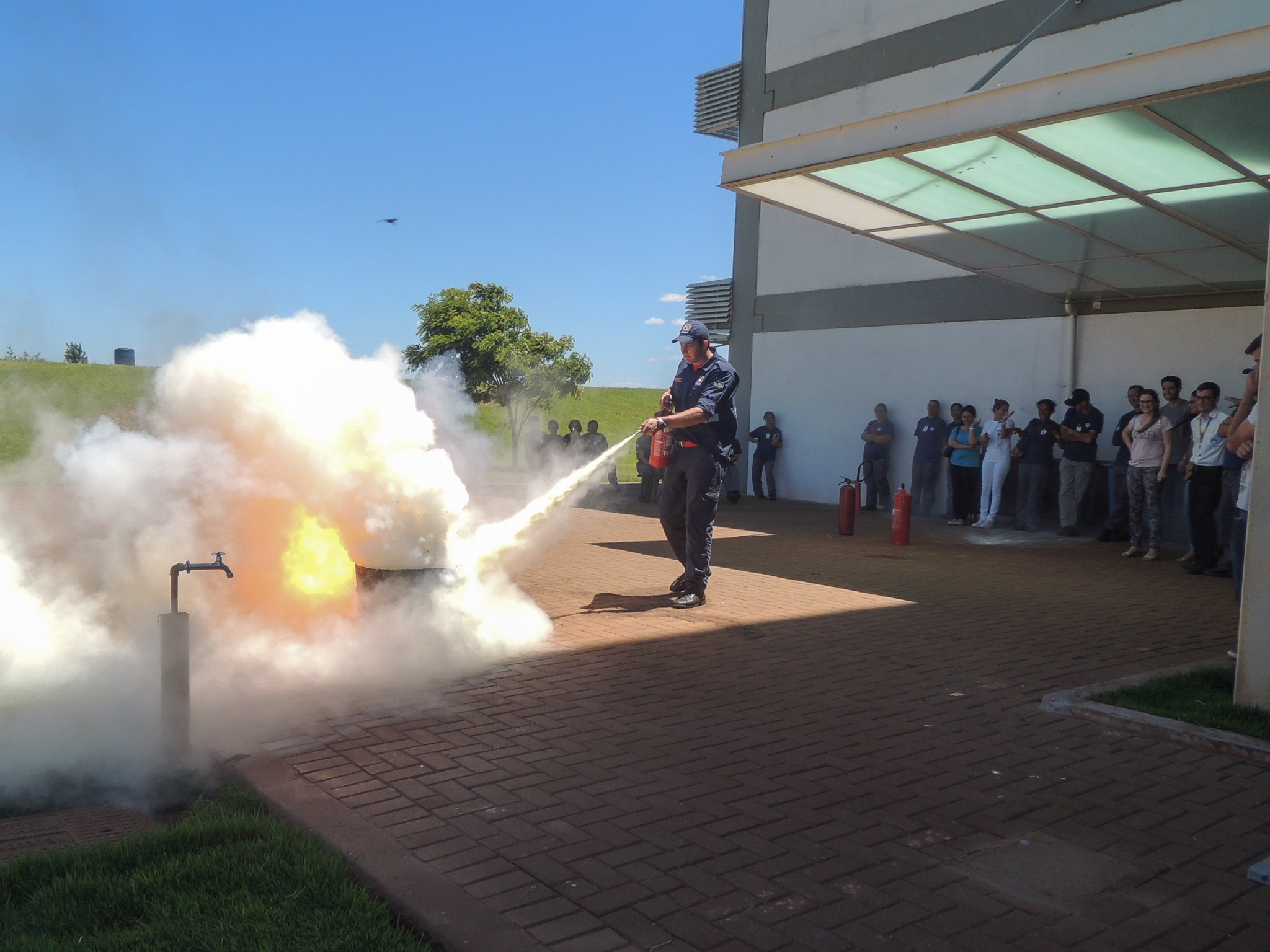 Do lado esquerdo, muita fumaça. Do lado direito, homem apagando fogo. Pessoas observando ao fundo.