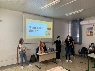 fotografia de três alunos de pé, na frente de uma sala de aula. No centro deles uma mesa de professor com um boneco de demonstração do corpo humano em cima. atrás um slide é projetado com o texto: O que é DM tipo II?