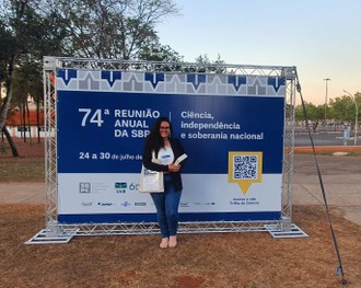 Uma menina posa de pé para a foto. Ela segura livros e uma bolsa nas mãos. Atrás dela um banner da Sociedade Brasileira para o progresso da ciência nas cores azul escuro e branco