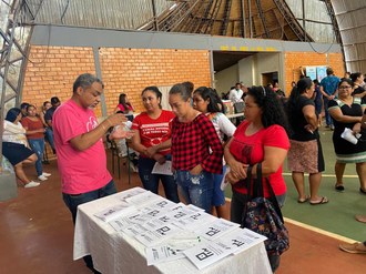 Um homem conversa com quatro mulheres. Todos estão de pé e do lado deles há uma mesa com algumas folhas de papel