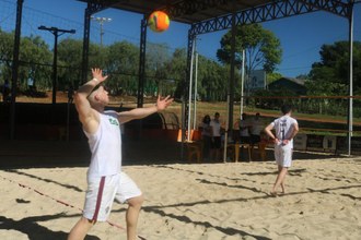 dois homens jogando vôlei de areia. Um aparece de costas em frente a rede e outro mais atrás sacando a bola