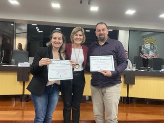 Duas mulheres  e um homem de pé posam para a foto.  Uma das mulheres e o homem seguram um papel nas mãos.