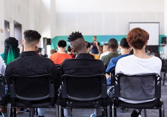 sala de aula. Tres estudantes aparecem sentados de costas. Ao fundo aparece um professor de pé.