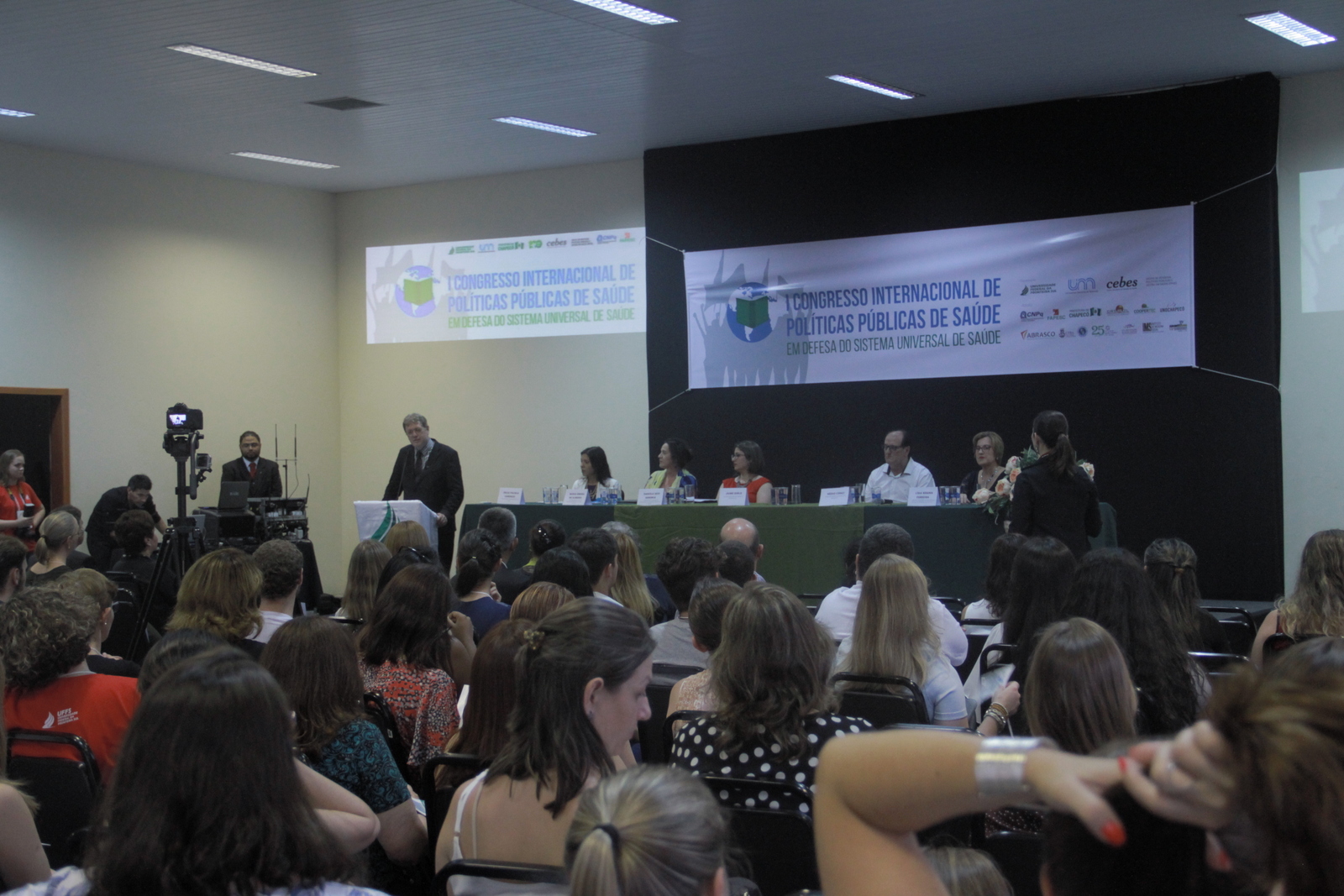 Diversas pessoas, sentadas, observam o palco principal do evento