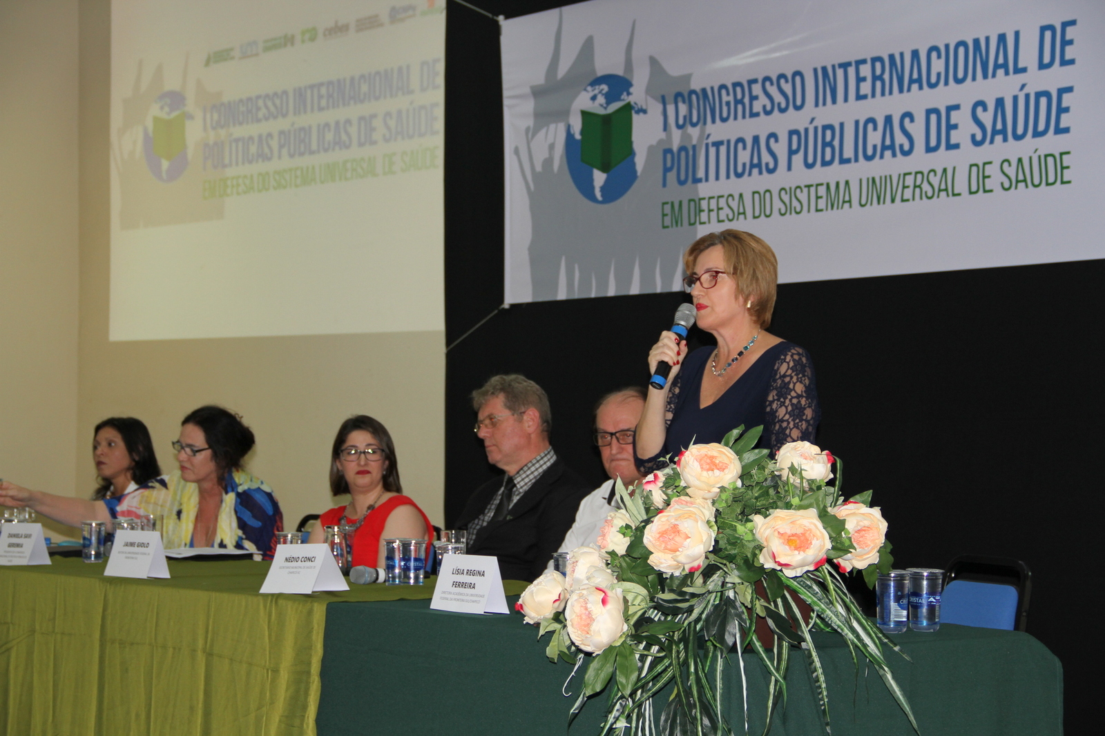 Mulher falando ao microfone na mesa oficial do evento