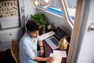 Um homem sentado na frente de uma mesa e escrevendo num caderno. Ao lado do caderno aparece um notebook aberto.